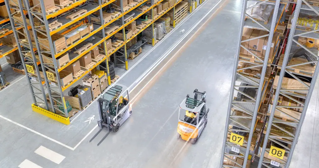Yüksek raflı bir depoda, raf koridorları arasında çalışan iki STILL RX 20 akülü forkliftin yukarıdan görünümü