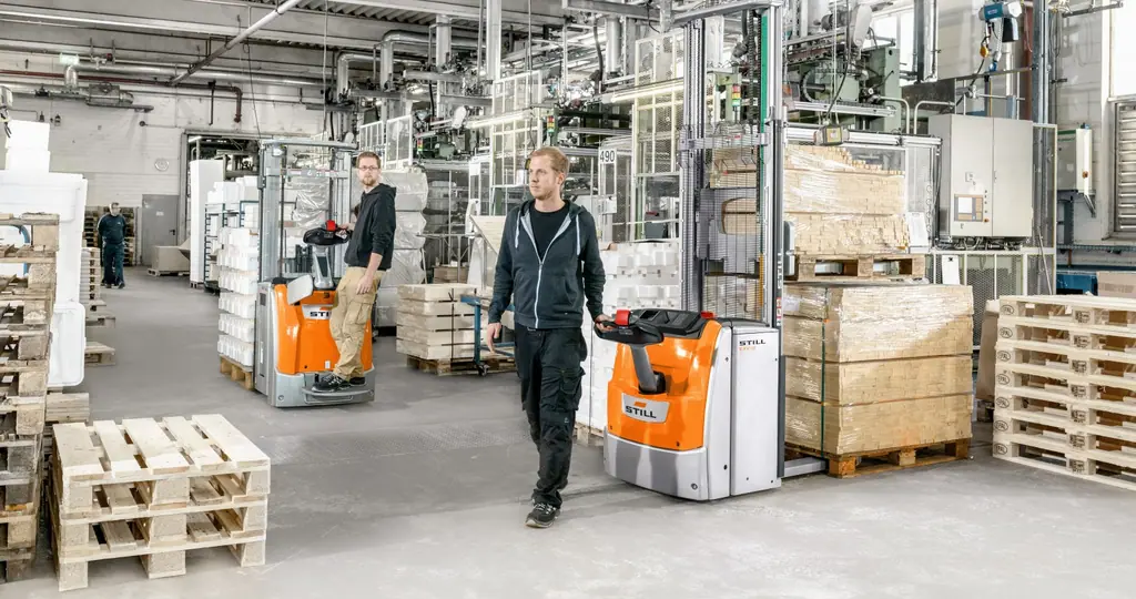Üretim alanında paletli yük taşıyan STILL EXV 14-20 serisi yaya kumandalı istif makinesi; arka planda ayakta kullanım platformlu bir STILL istif makinesi