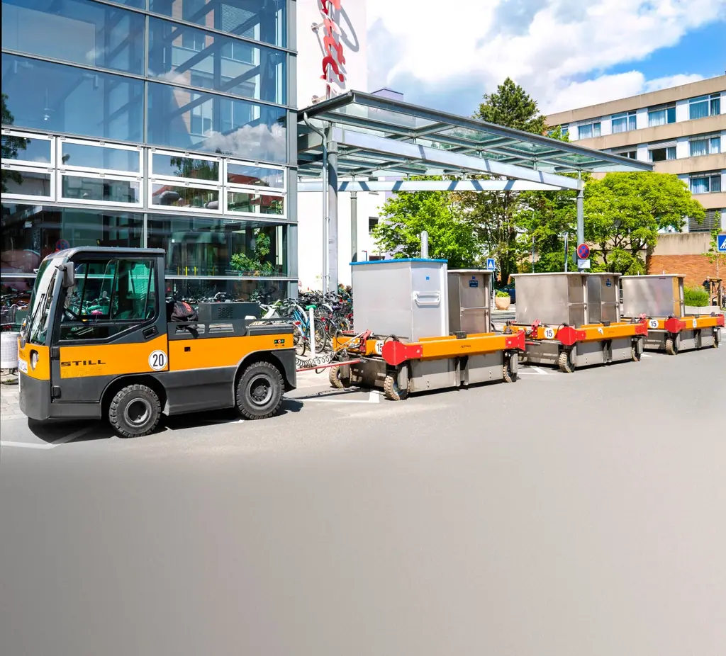 STILL R 07-25 çekici ve üç LiftRunner C-çerçeve römorktan oluşan çeki treni bina girişinde bekliyor (STILL referans fotoğrafı; alt kısmı metin alanı için rötuşlanmış gri zemin, kırpılması önerilir)