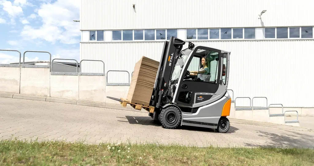 STILL RX 60-25/35 serisi akülü forklift, endüstriyel bina önündeki eğimli yolda karton yüklü paletle ilerlerken