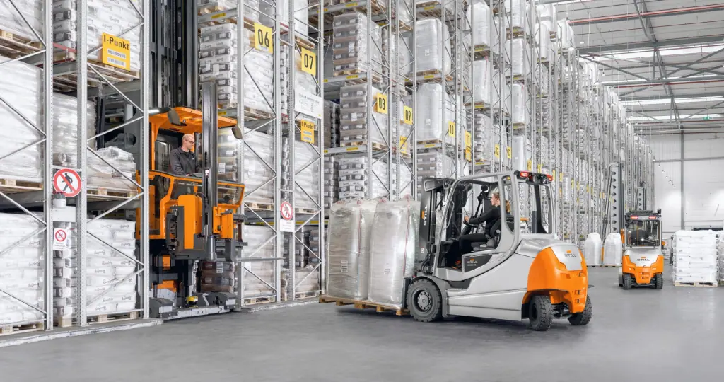 STILL RX 60-35/50 serisi forklift, yüksek raflı depoda big-bag paleti taşırken; solda dar koridor istif aracı